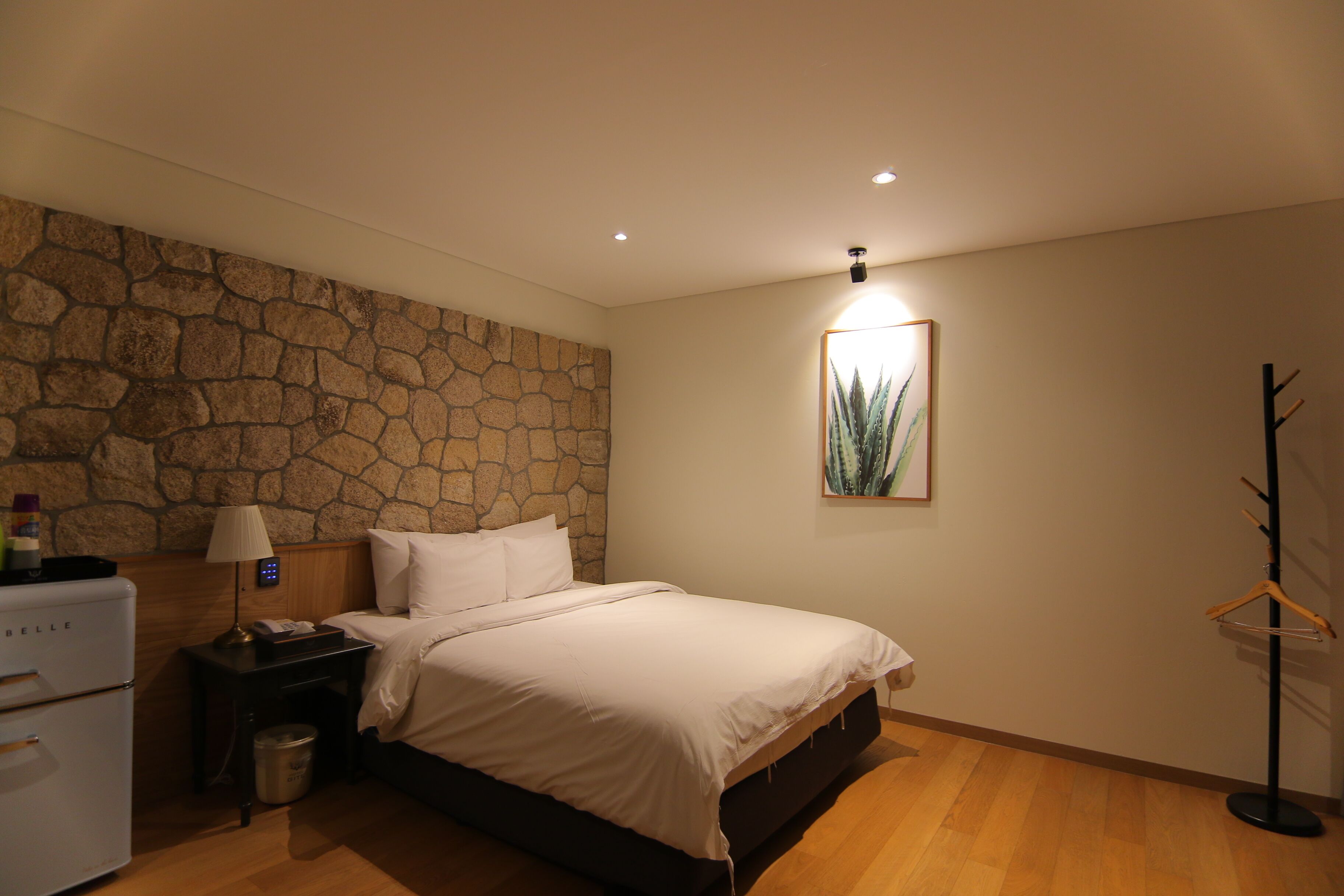 A bedroom with a stone wall, a bed, a nightstand with a lamp, a coat rack, and a small refrigerator.