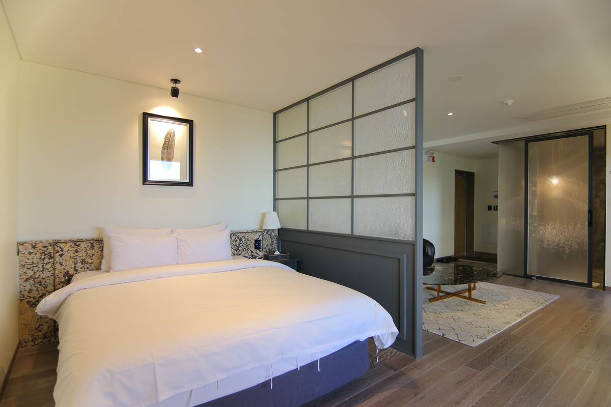 Una habitación de hotel moderna con una cama grande, una mesita de noche, un cuadro enmarcado en la pared y una puerta corrediza de vidrio que da a un baño.
