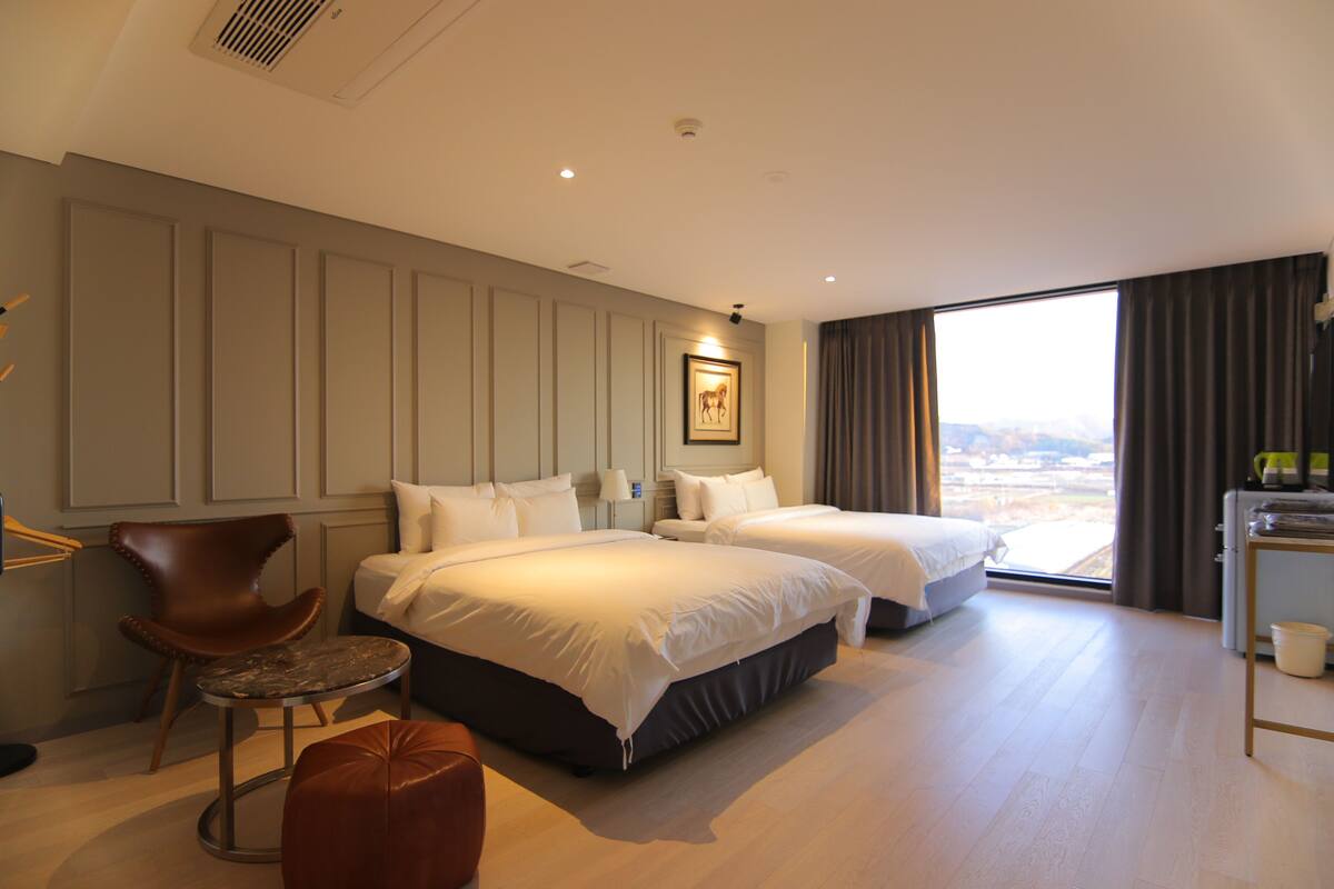 Habitación de hotel con dos camas, una silla, una mesita y vistas a la ciudad.