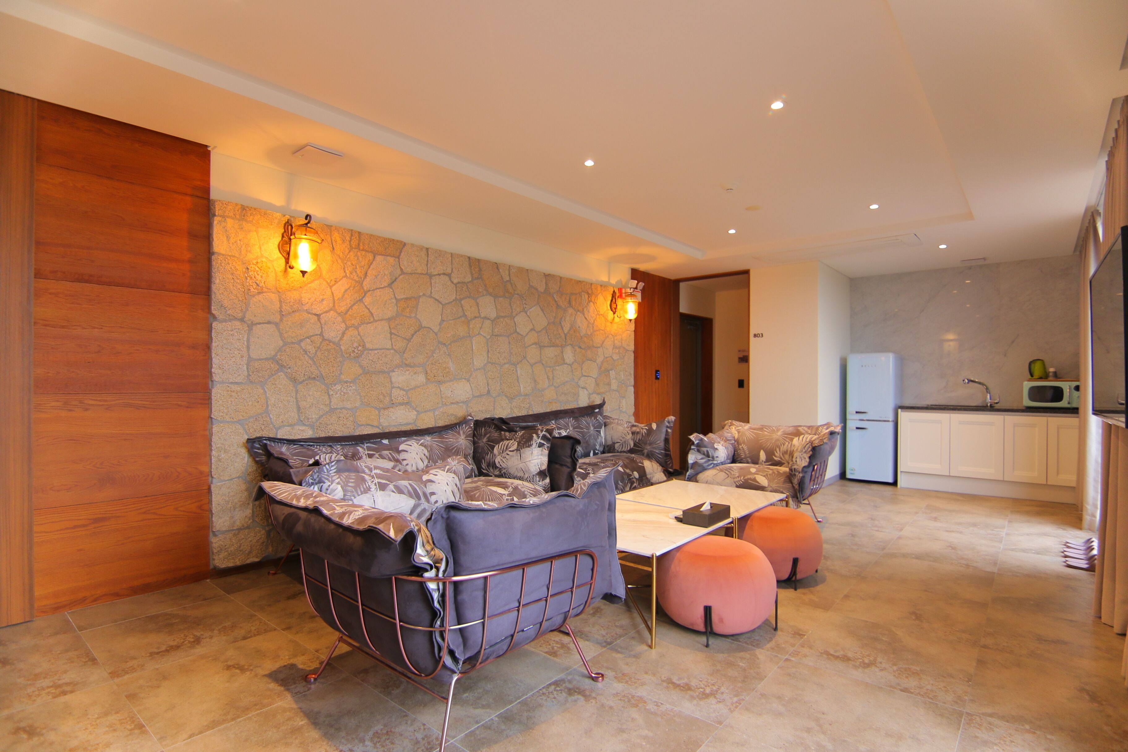 Un salón moderno con pared de piedra, panel de madera, sofá con cojines, una mesa de centro y una cocina al fondo.