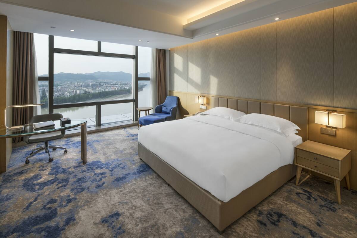 A hotel room with a large bed, a desk, a chair, and a view of a cityscape through large windows.
