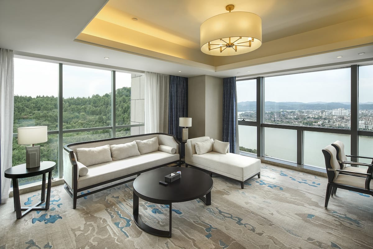 A spacious living room with a sofa, armchair, and coffee table, large windows with a city view, and a modern chandelier.