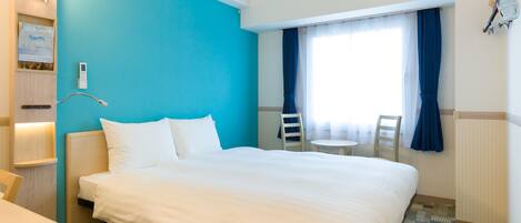 A hotel room with a large bed, a bedside table, a small desk, a chair, a window with curtains, and a wall-mounted lamp.