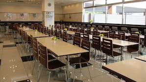 Eine großzügige Cafeteria mit mehreren Tischen und Stühlen, großen Fenstern und einer Uhr an der Wand.
