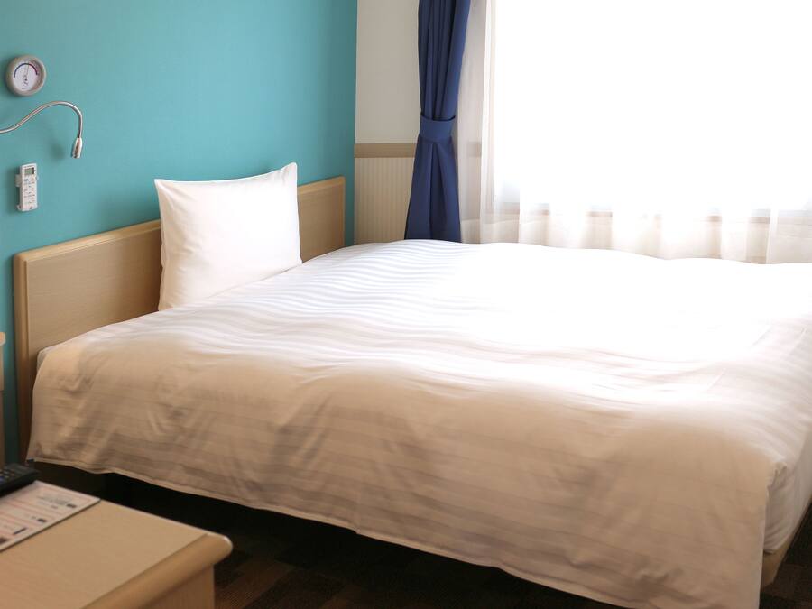 A hotel room with a bed, a bedside table, a clock, and a window with curtains.