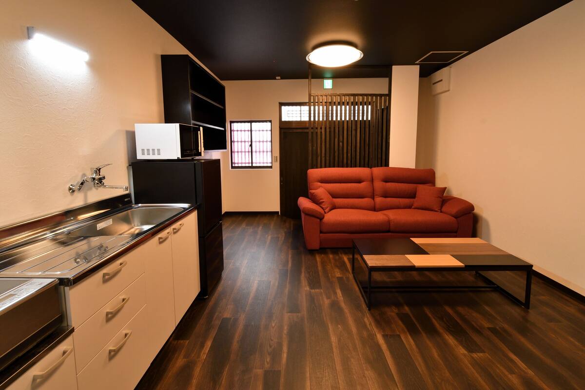 A compact kitchen with modern appliances, a wooden floor, a red sofa, and a small table.