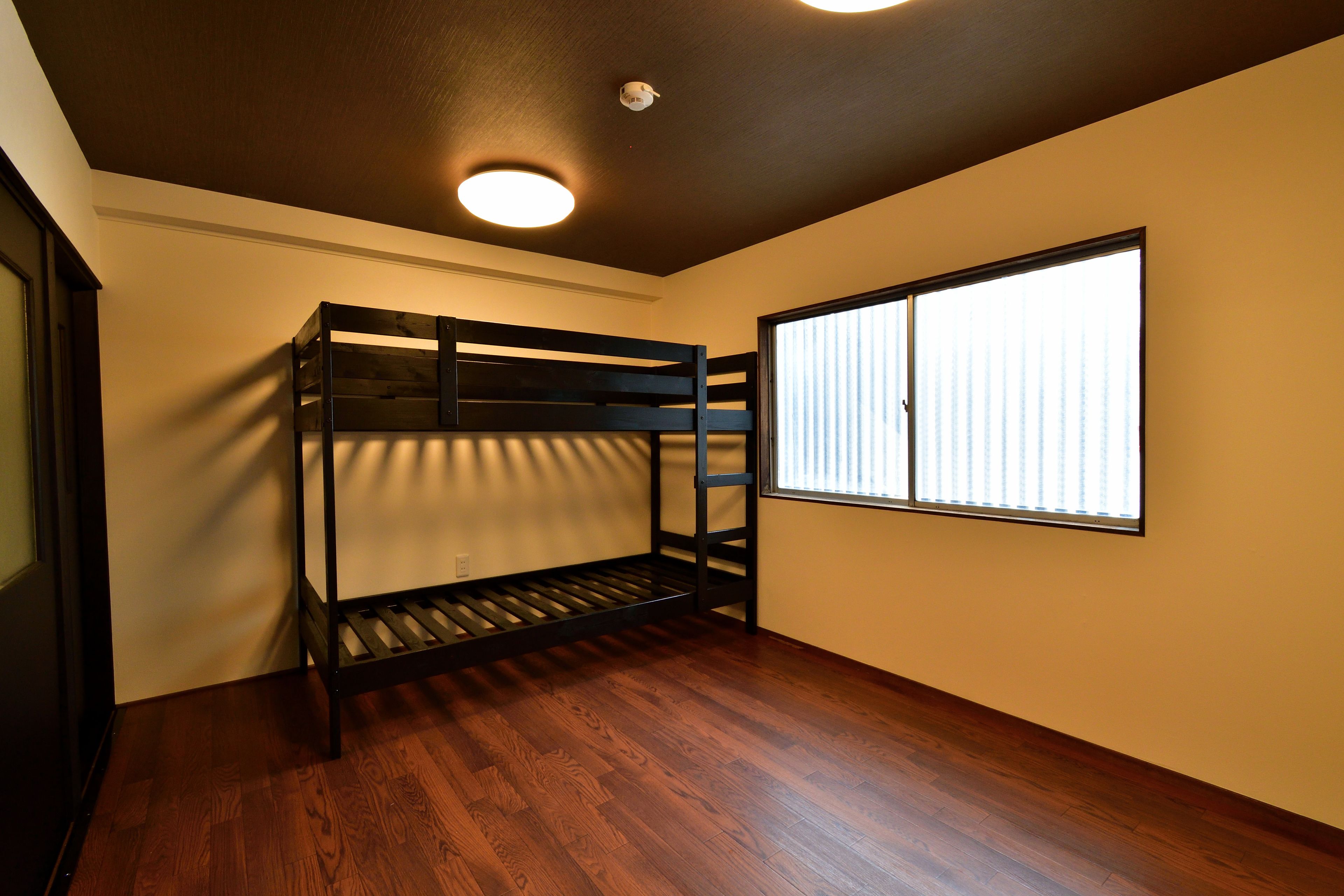 A room with a bunk bed, wooden flooring, a window with blinds, and a ceiling light.