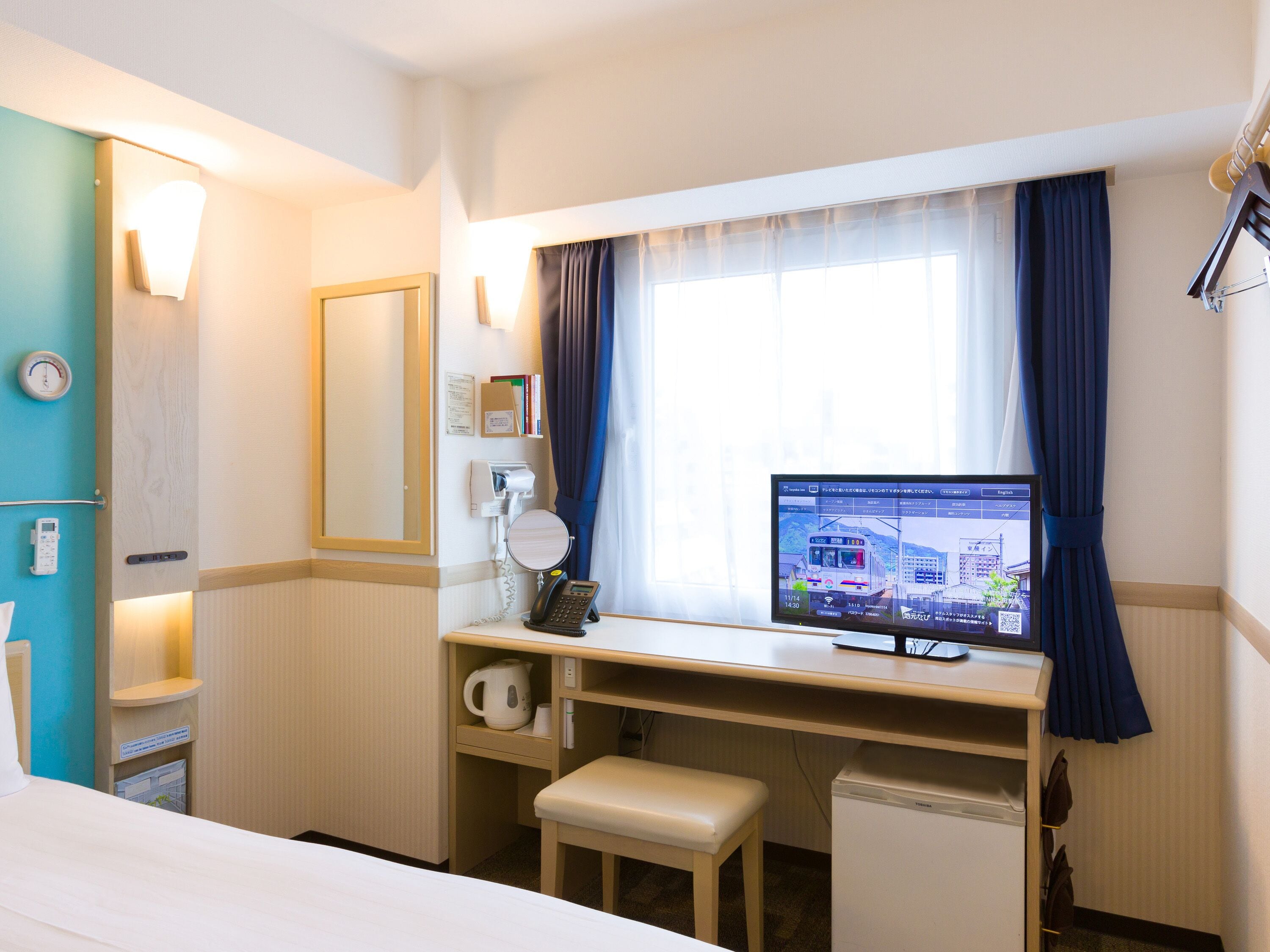 A hotel room with a bed, a desk with a computer monitor displaying a train schedule, a telephone, and a small cabinet.