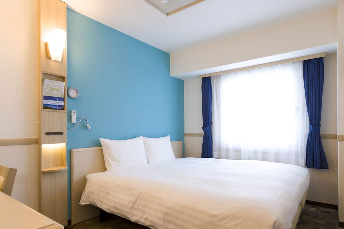 A hotel room with a large bed, a desk, a chair, a wall clock, a thermostat, and a window with curtains.