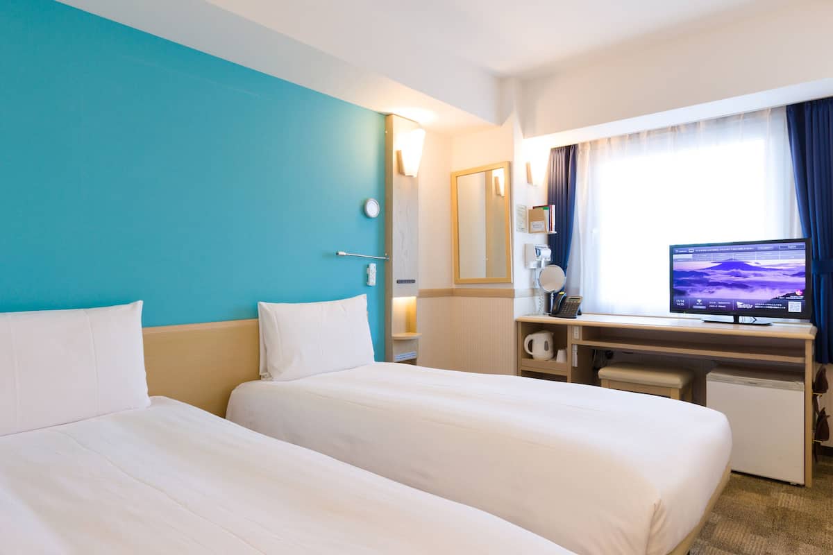 A hotel room with two beds, a desk with a computer, a coffee maker, and a window with curtains.