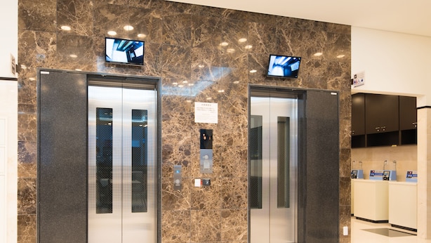 Deux ascenseurs équipés d’écrans numériques, un mur en marbre et un espace d’accueil avec un comptoir et des panneaux d’information.