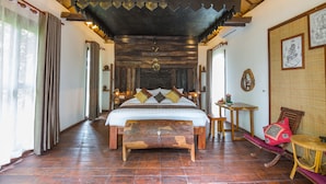 Une chambre spacieuse avec un grand lit, une tête de lit en bois et un ventilateur de plafond. On y trouve une petite table et une chaise dans un coin.