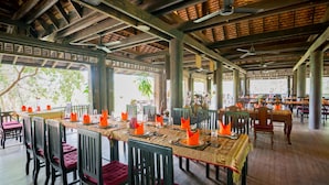 Une salle à manger spacieuse, dotée de hauts plafonds et de poutres en bois, où les tables sont dressées avec des serviettes orange et des coussins rouges.