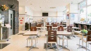 A spacious dining area with multiple tables and chairs, a modern kitchen in the background, and large windows providing natural light.