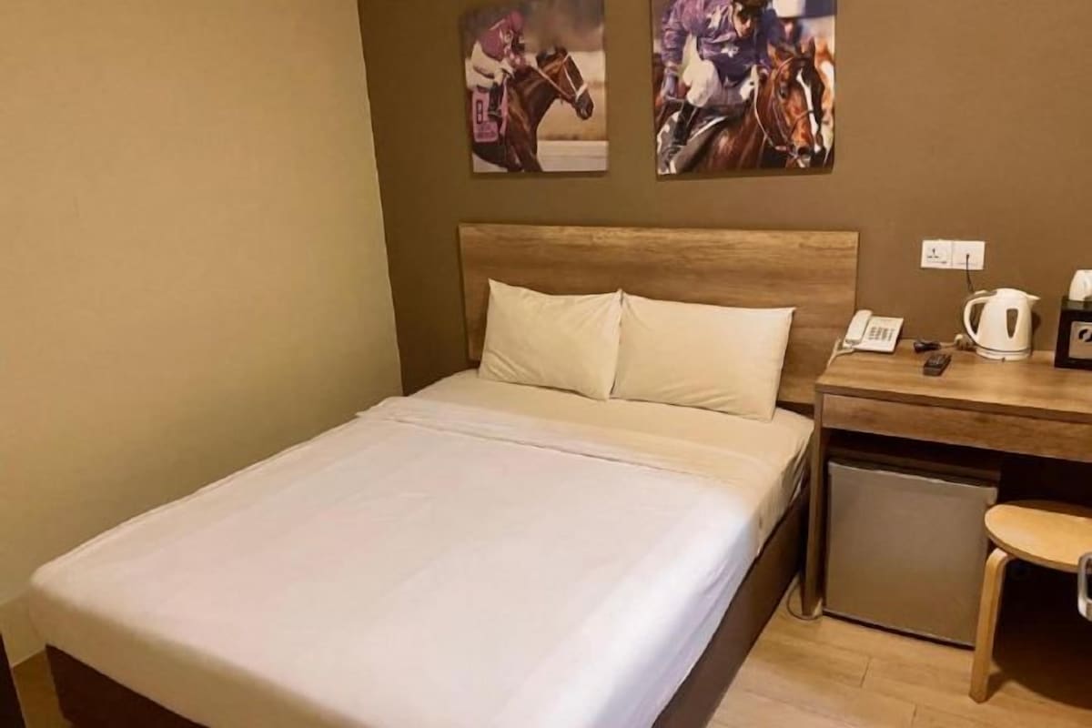Une chambre d’hôtel avec un lit, une tête de lit en bois, un petit bureau avec un téléphone et une bouilloire, et une prise électrique fixée au mur.