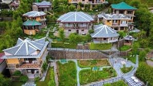 A residential area with houses featuring sloped roofs and balconies, surrounded by lush greenery and a stone wall. - Udechee Huts Hotel (Dharamshala)