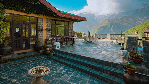 A building with a stone facade, large windows, and a red roof. There are steps leading to a terrace with outdoor seating and potted plants. - Udechee Huts Hotel (Dharamshala)
