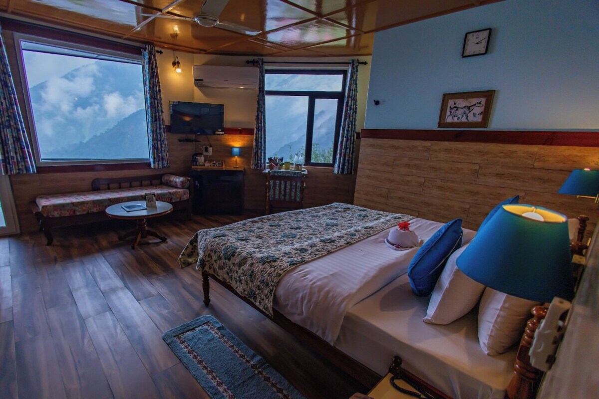 A bedroom with a bed, bedside lamps, a desk, a chair, a small table, and a window with a view of mountains.