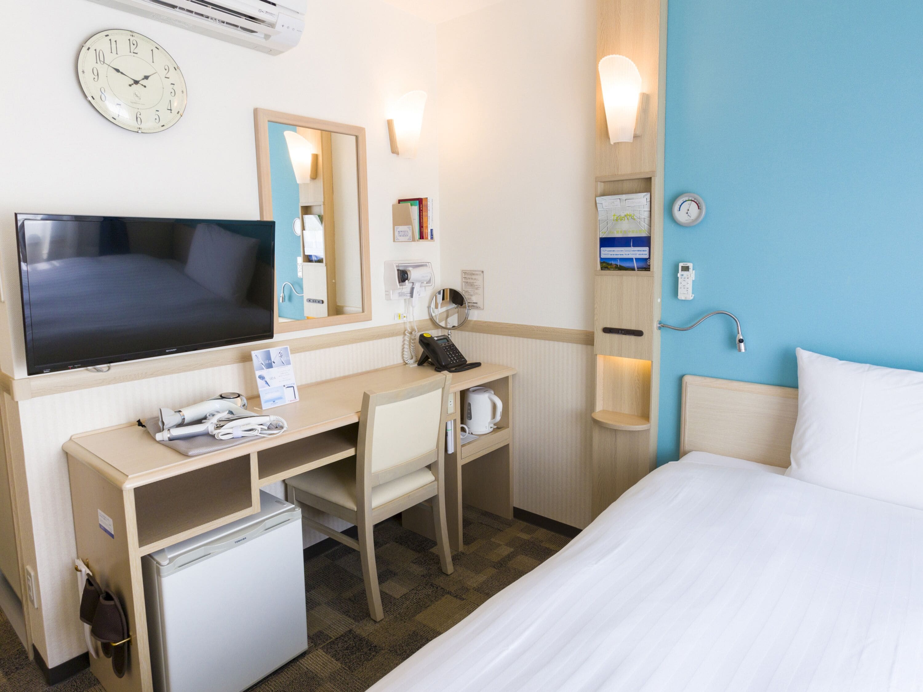Une chambre d’hôtel équipée d’un lit, d’un bureau avec un ordinateur, d’une télévision, d’un réfrigérateur, d’un four à micro-ondes, d’une machine à café, d’une horloge et d’un miroir.