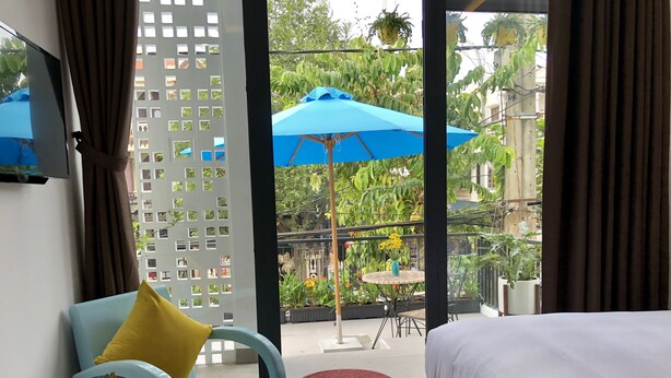 A hotel room with a bed, a blue chair with a yellow pillow, a small table with a fruit bowl, and a view of an outdoor area with a blue umbrella and plants. - La An Homestay (Da Nang)