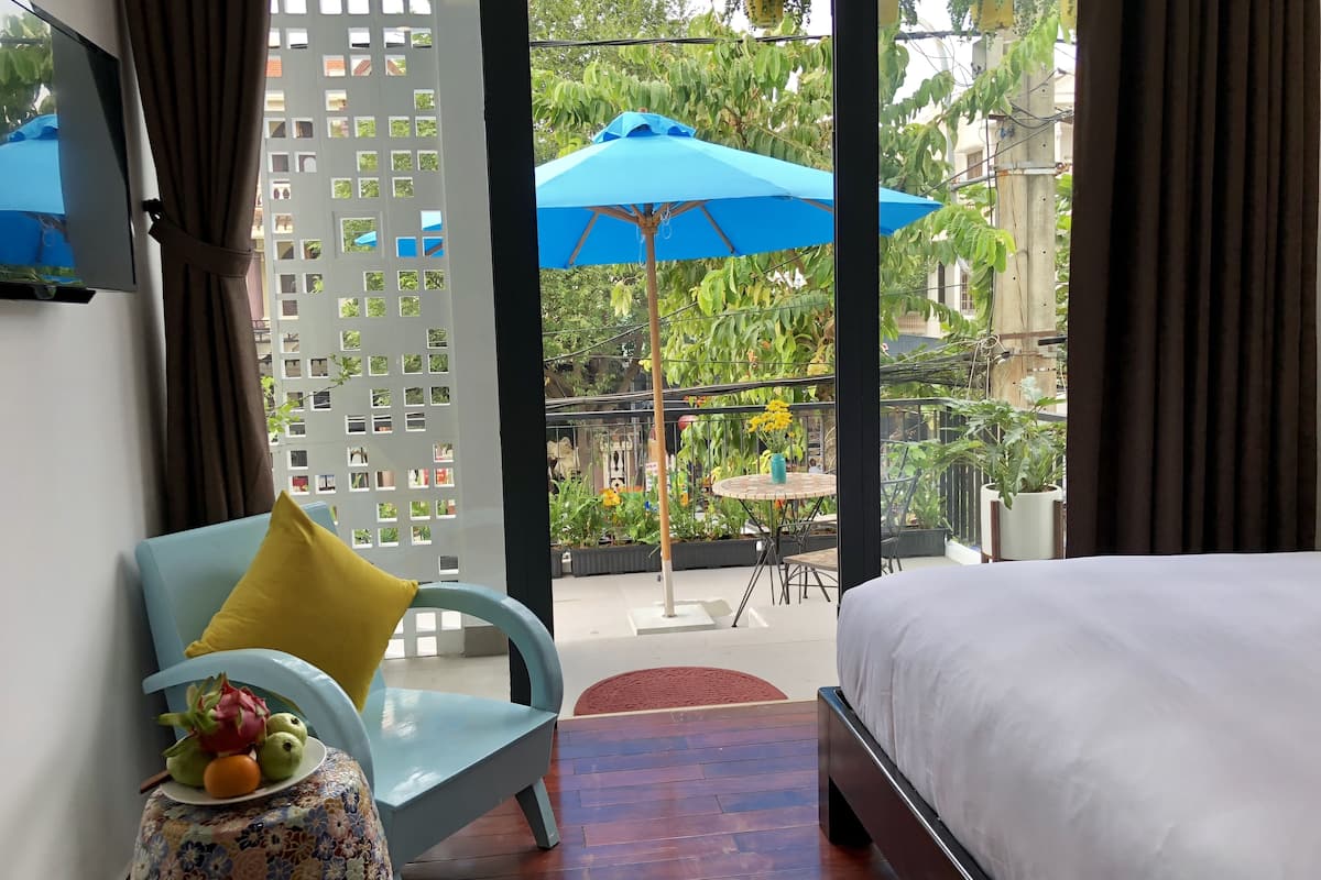 A hotel room with a bed, a blue chair with a yellow pillow, a small table with a fruit bowl, and a view of an outdoor area with a blue umbrella and plants.