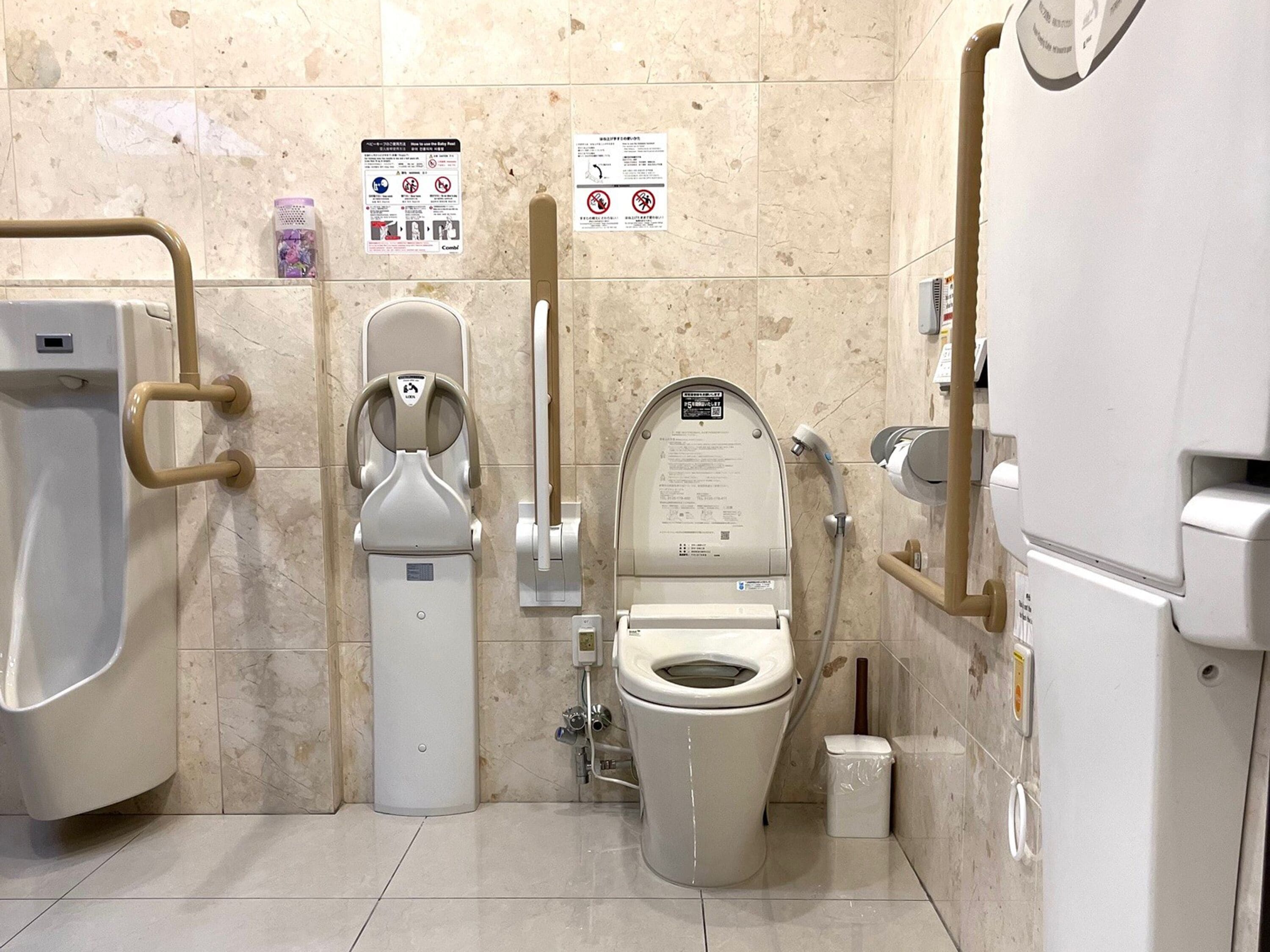 Des toilettes pour hommes comprenant un urinoir, des toilettes équipées d’un siège-bidet et un distributeur de gel hydroalcoolique.