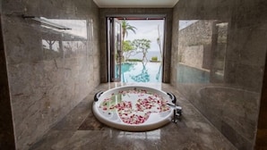 A modern bathroom with a large bathtub filled with rose petals, marble walls, and a view of a pool and greenery. - Villa Maiara (Ungasan)