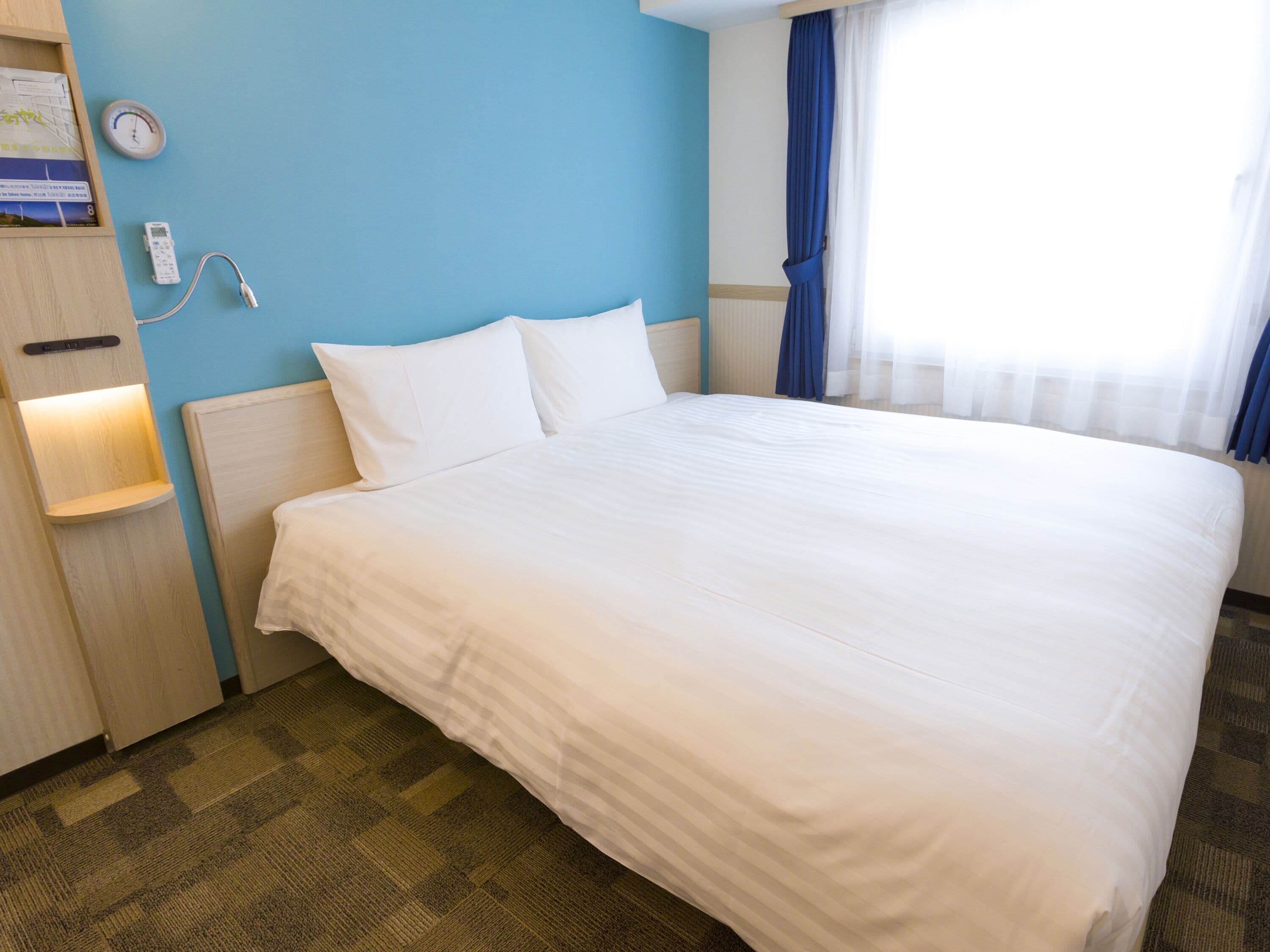 A hotel room with a large bed, a nightstand, a wall clock, and a window with curtains.