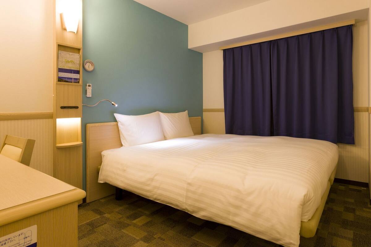 Une chambre d’hôtel avec un lit, un bureau, une table de chevet, une horloge murale et une fenêtre avec des rideaux.
