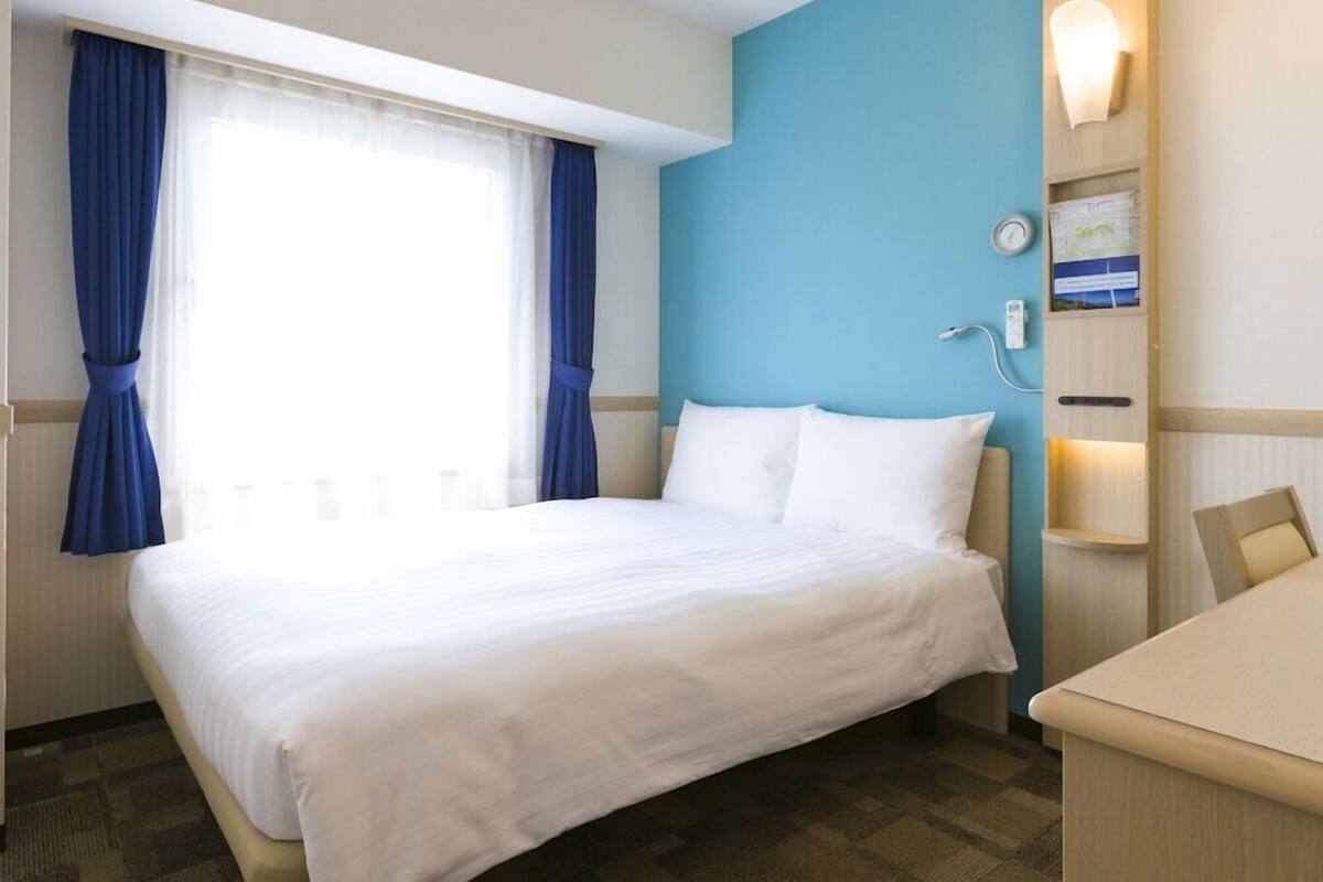 Une chambre d’hôtel comprenant un lit, un bureau, une fenêtre avec des rideaux, une horloge murale et une étagère avec un téléphone et une carte.