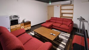 A furnished room with a red sofa, a wooden coffee table, a microwave, a small cabinet, and a shelf with books. - Machi de kurasu Tengachaya-2 (Osaka)