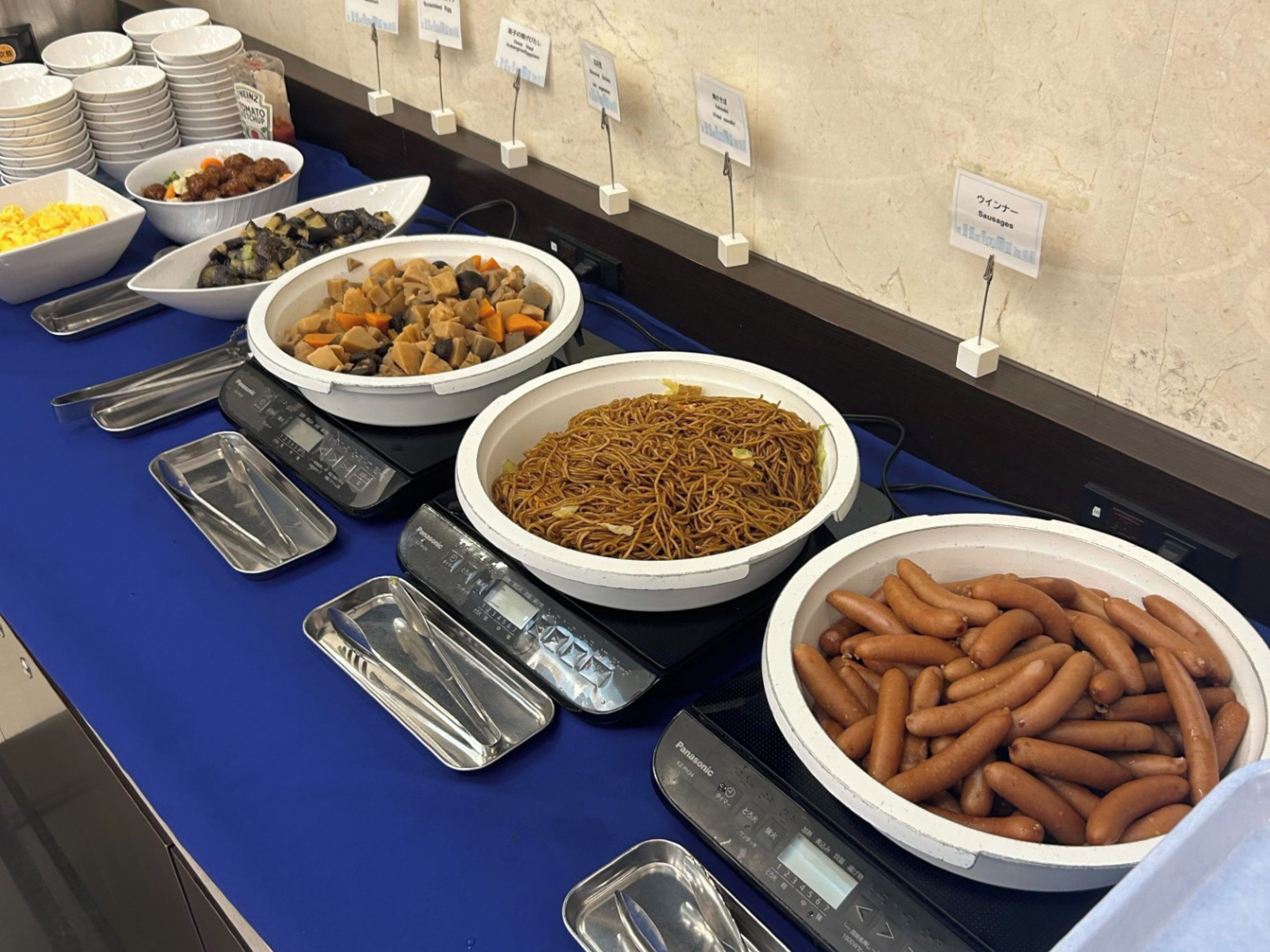 En buffet med forskellige retter, herunder pølser, nudler og grøntsager.