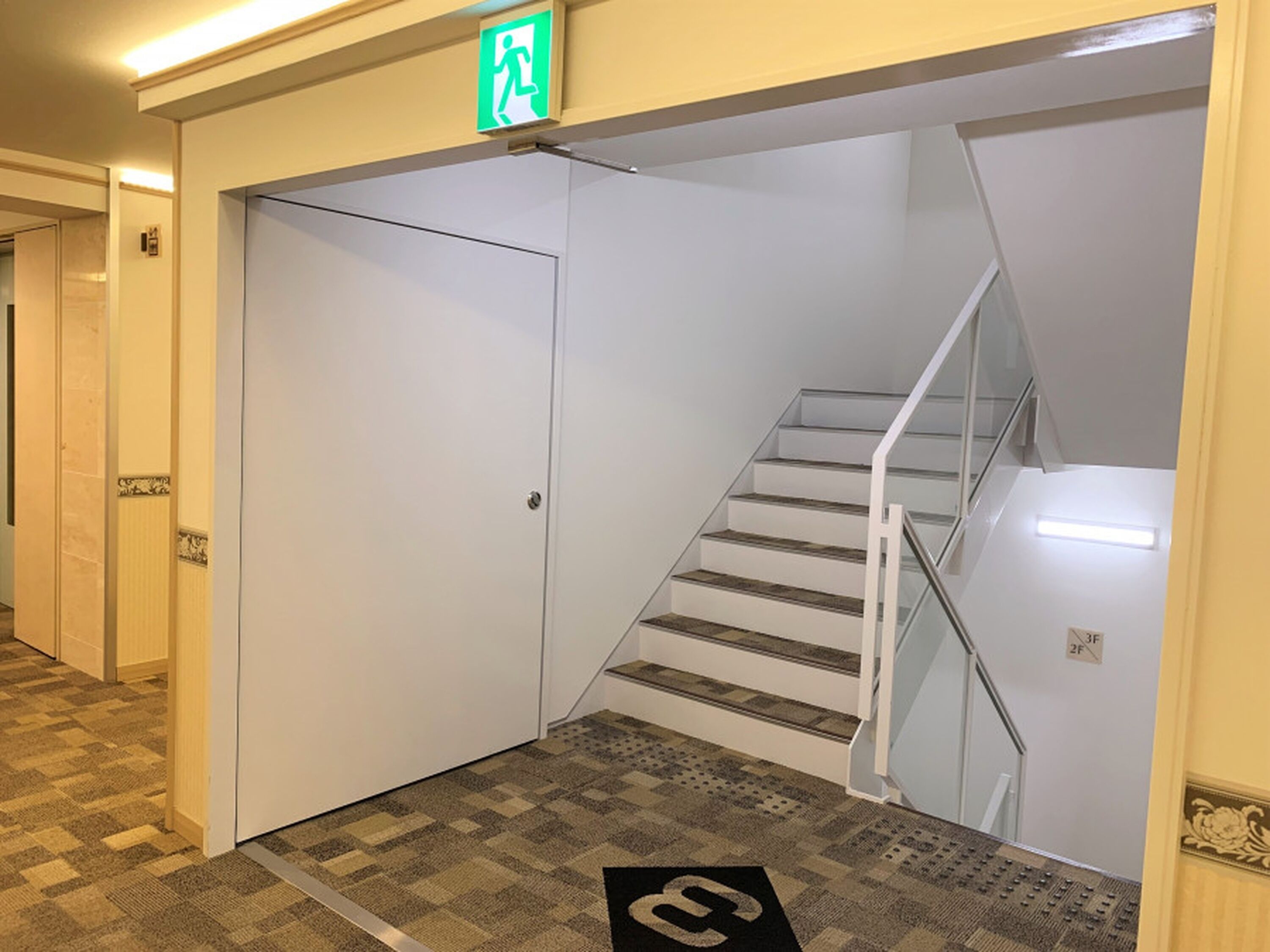 A hotel interior with a staircase, a door with an emergency exit sign, and a patterned carpet.