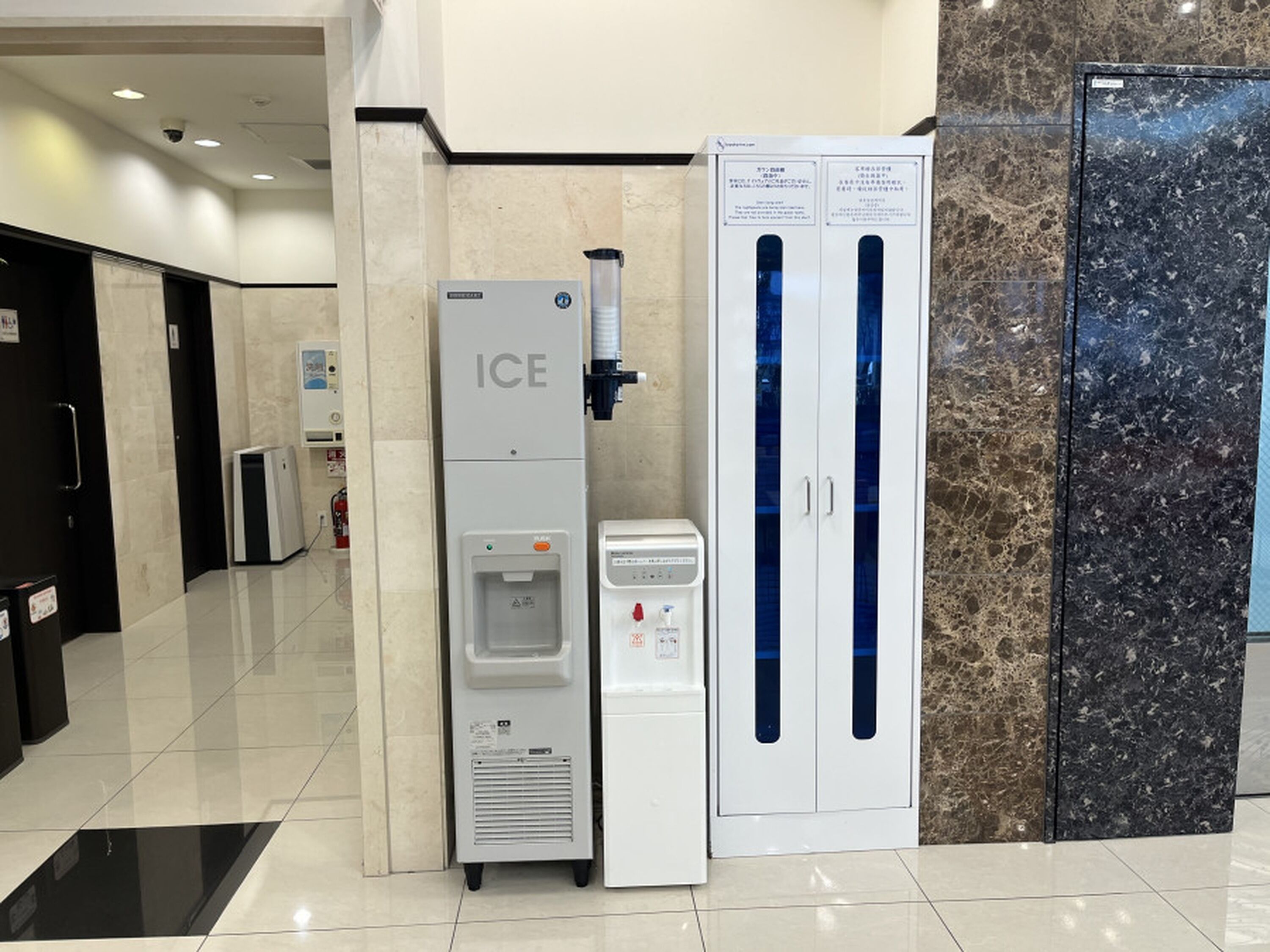A hotel lobby with a water dispenser, a refrigerator, and a marble wall.
