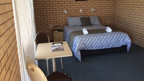 Un lit simple avec une couvre-lit à motifs, une petite table avec une télécommande, une chaise et un tableau représentant une orchidée, accroché au mur.