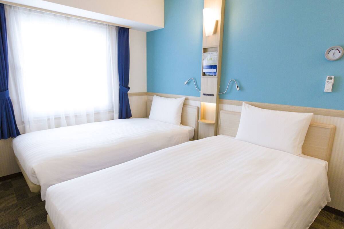 Ein Hotelzimmer mit zwei Betten, einem Fenster mit Vorhängen, einer Wanduhr und einem Nachttisch mit Lampe.