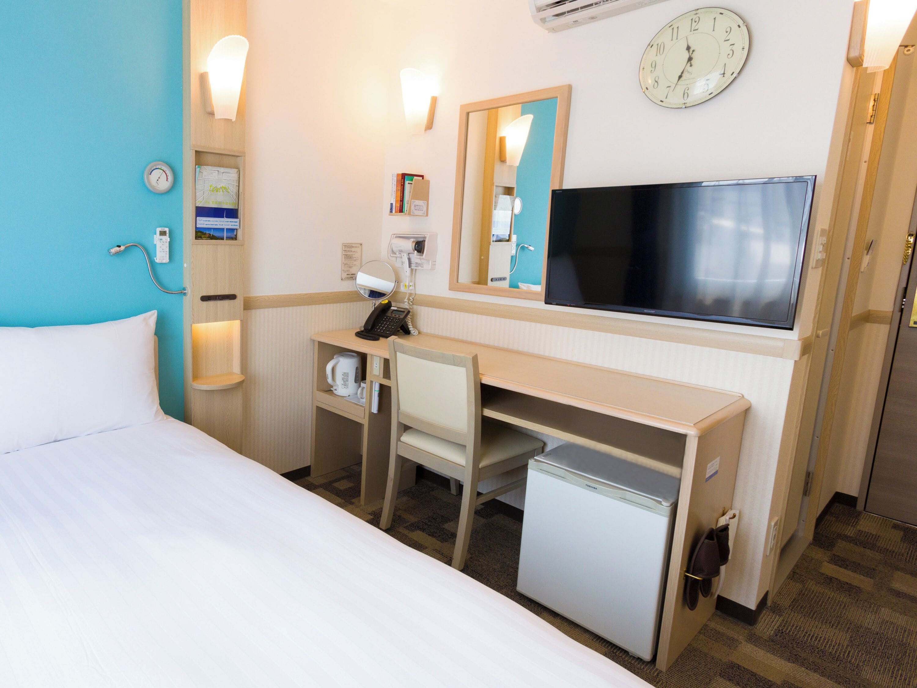 Une chambre d’hôtel équipée d’un lit, d’un bureau, d’une chaise, d’un petit réfrigérateur, d’une télévision, d’une horloge et d’un miroir.
