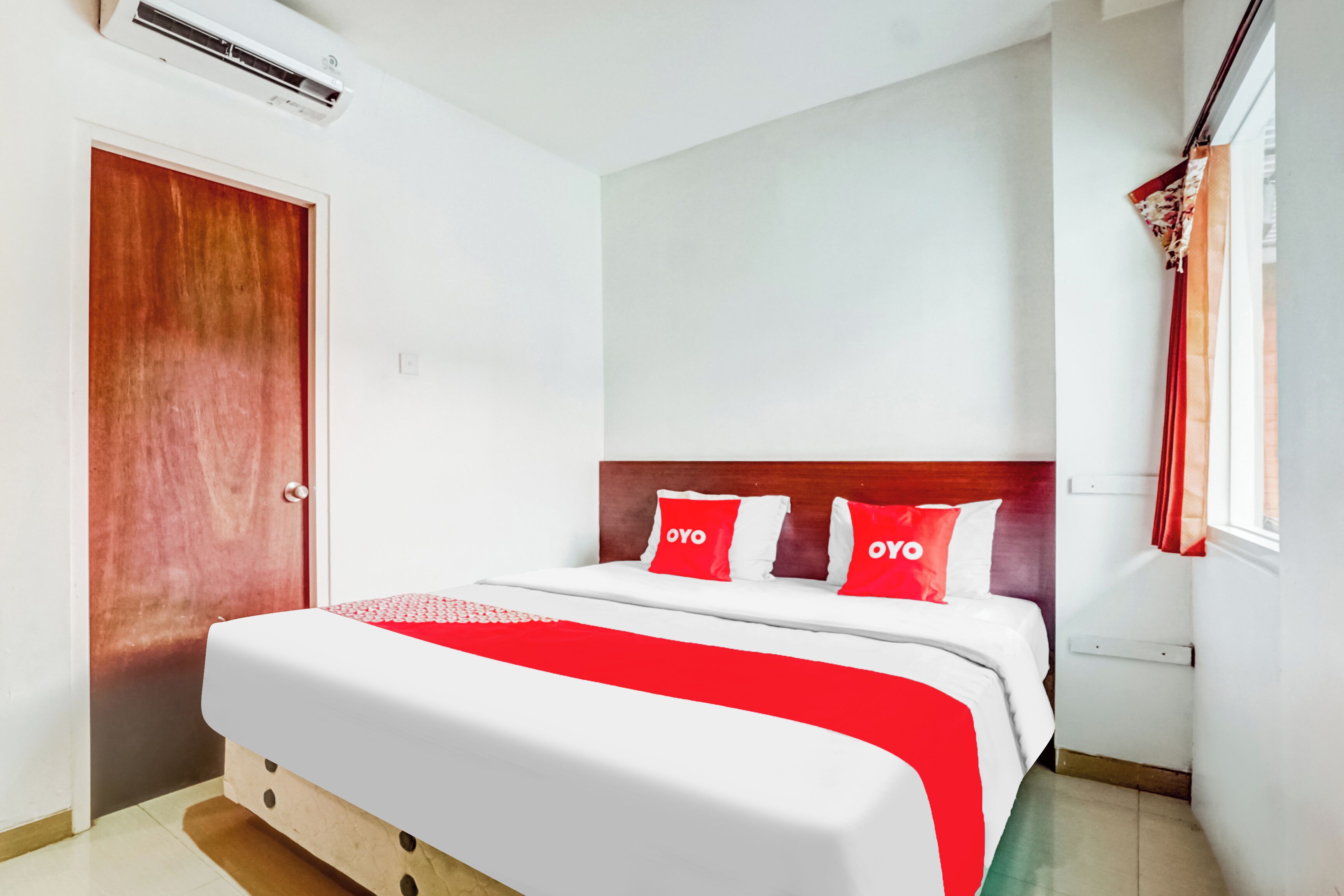 Une chambre d’hôtel avec une porte en bois, un lit avec une literie blanche et rouge, et une fenêtre avec des rideaux rouges.