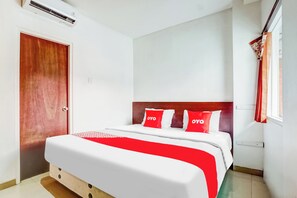 A hotel room with a wooden door, a bed with white and red bedding, and a window with red curtains.