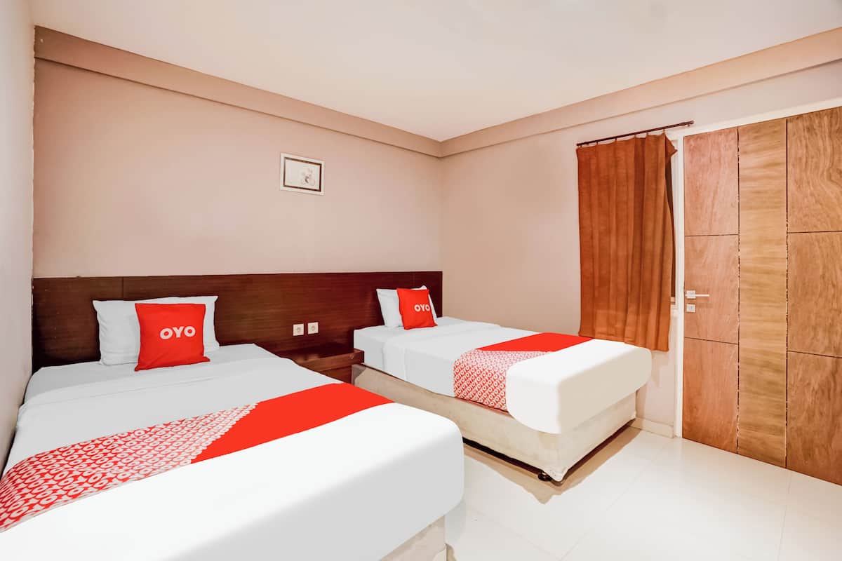 Quarto de hotel com duas camas, cada uma com roupa de cama branca e detalhes vermelhos, cabeceira de madeira, guarda-roupa de madeira e um pequeno quadro emoldurado na parede.