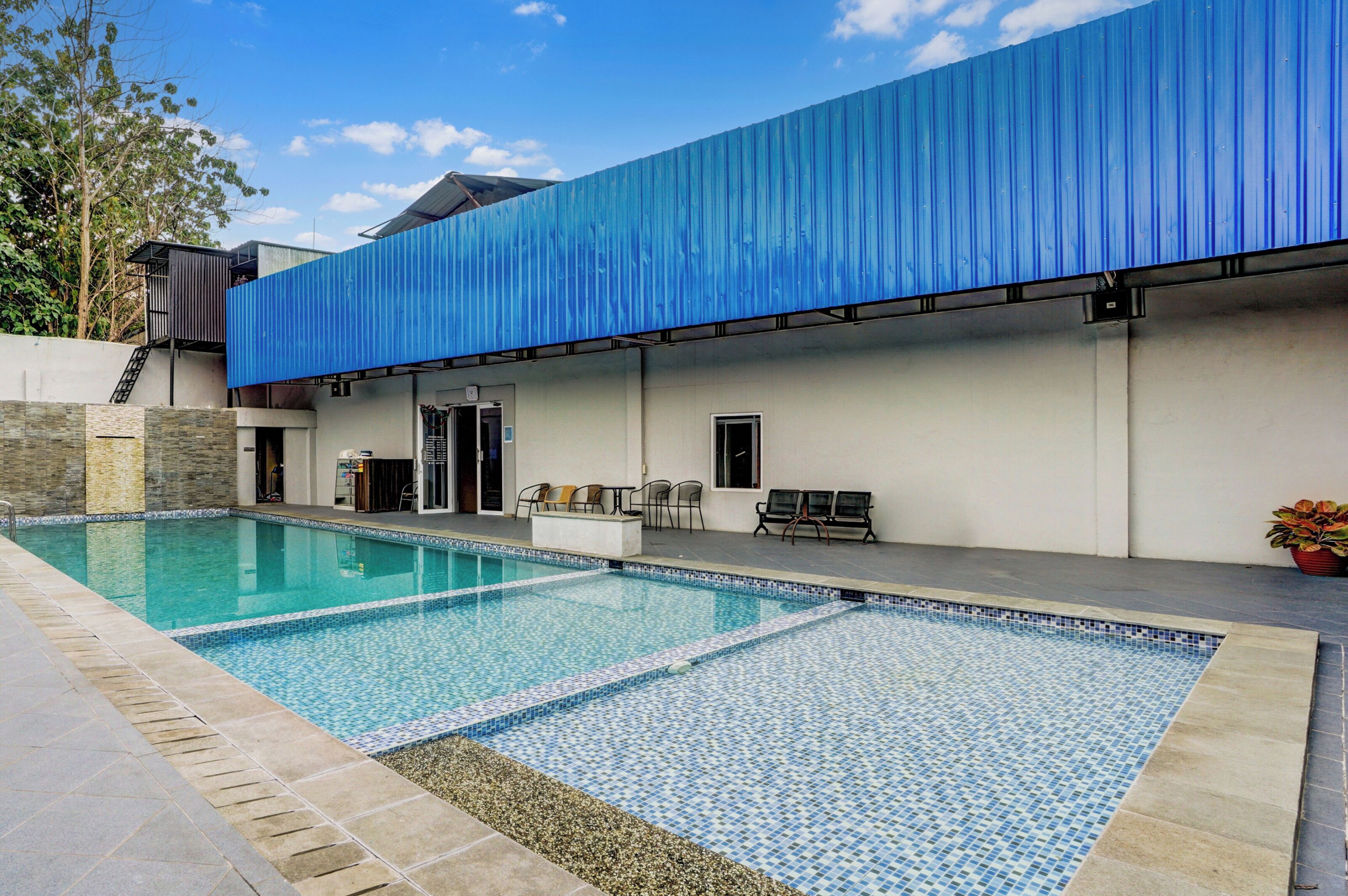 A swimming pool with a tiled deck, a covered patio area, and a building with a blue exterior.