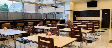 A modern dining area with multiple tables and chairs, large windows, and a kitchen area in the background.