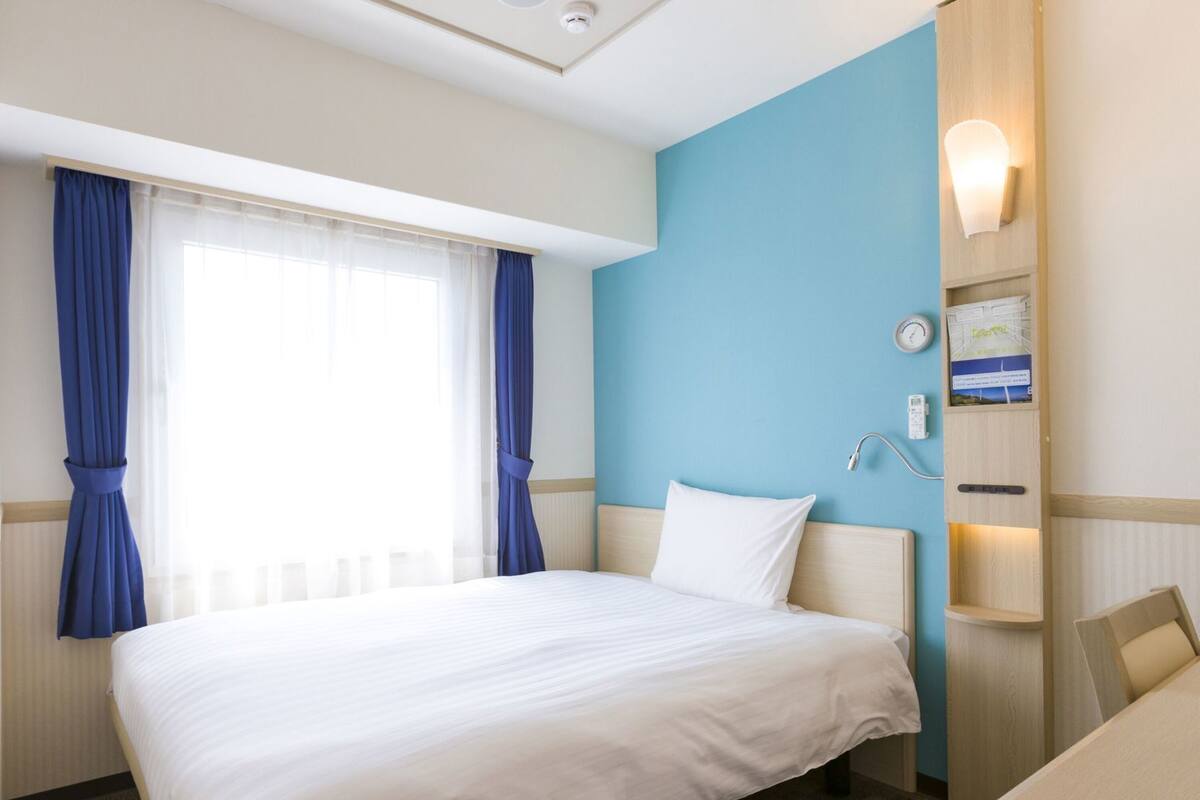 Een hotelkamer met een bed, bureau, stoel, raam met gordijnen, een wandklok en een plank met een boek en een afstandsbediening.