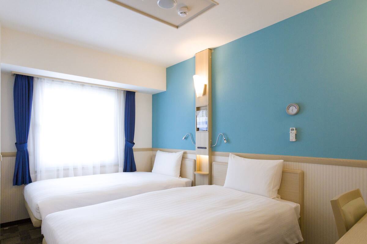 Een hotelkamer met twee bedden, een groot raam met gordijnen, een wandheater en een klok.