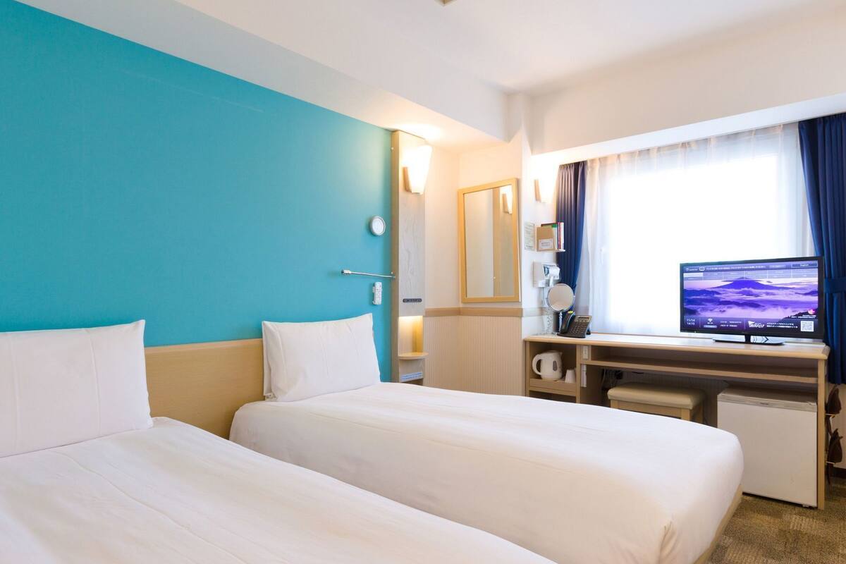 Een hotelkamer met twee bedden, een bureau met een computer, een kleine koelkast en een raam met gordijnen.