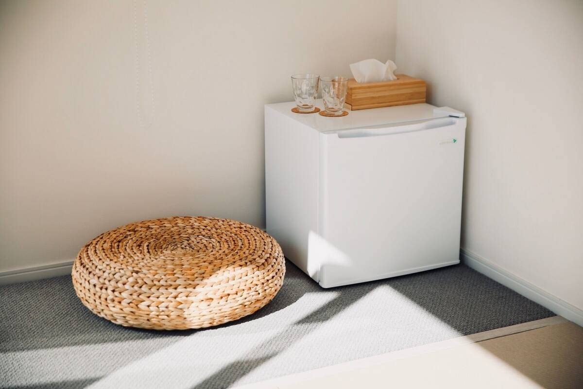 A corner of a room with a white refrigerator, a woven round ottoman, and a wooden box with tissues on top.