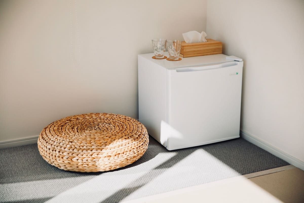 Esquina de una habitación con un refrigerador blanco, un otomán redondo tejido y una pequeña caja de madera con pañuelos encima.