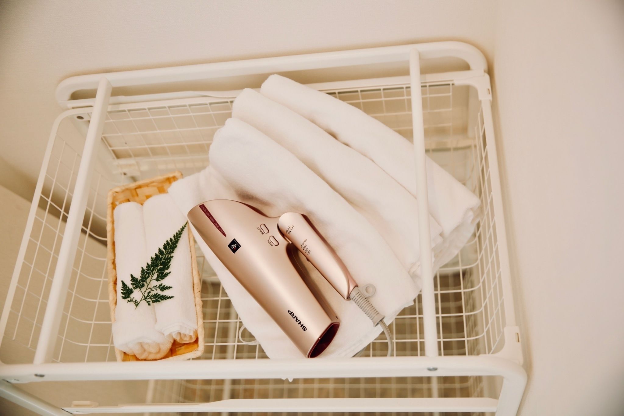 Een witte draadmand met opgevouwen handdoeken, een haardroger en een klein handdoekje met een varenblad.