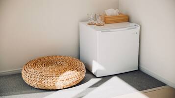 Una esquina de una habitación con un refrigerador blanco, un otoman tejido redondo y una caja de madera con pañuelos encima.