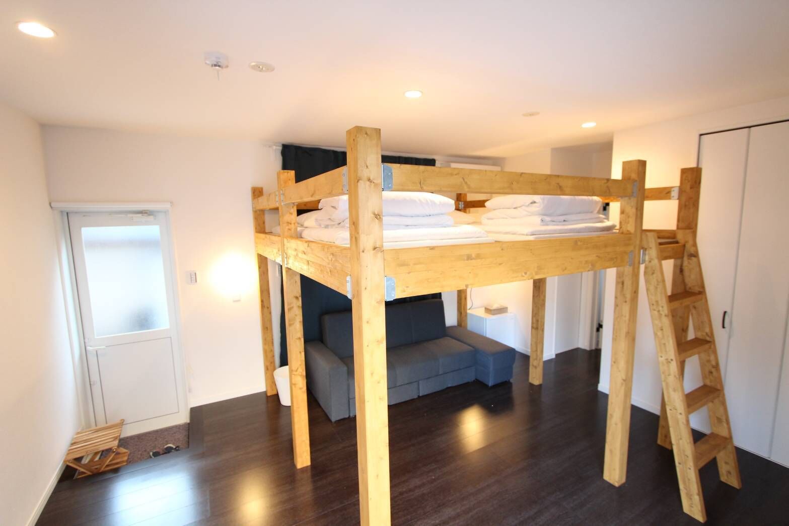 A bunk bed with a ladder, a sofa, and a wardrobe in a room with wooden flooring and white walls.